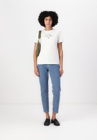 Hvid kortærmet t-shirt med broderet logo, parret med lyseblå skinny jeans og sorte loafers, båret med en grøn skuldertaske.