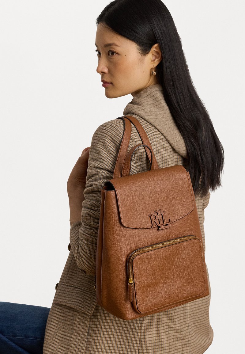 Mochila de couro castanho com um formato estruturado, apresentando ferragens douradas e um bolso exterior com fecho zip. Logótipo em relevo na aba.