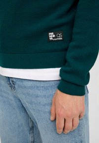 Strickpullover in strukturiertem Dunkelgrün mit gerippten Bündchen, versehen mit einem kleinen Markenlabel, über einem weißen Hemd getragen, kombiniert mit hellblauen Jeans.