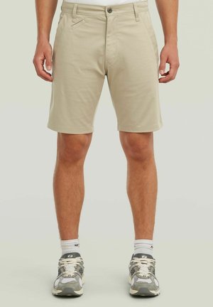 Mann trägt beige knielange Shorts, weiße Socken und graue Laufschuhe, steht vor einem einfarbigen hellen Hintergrund.