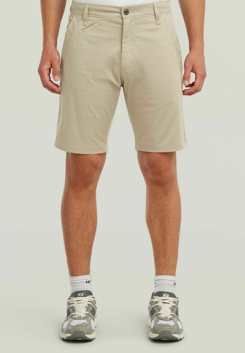 Homme portant un short beige au genou, des chaussettes blanches et des chaussures de course grises, debout devant un fond clair uni.