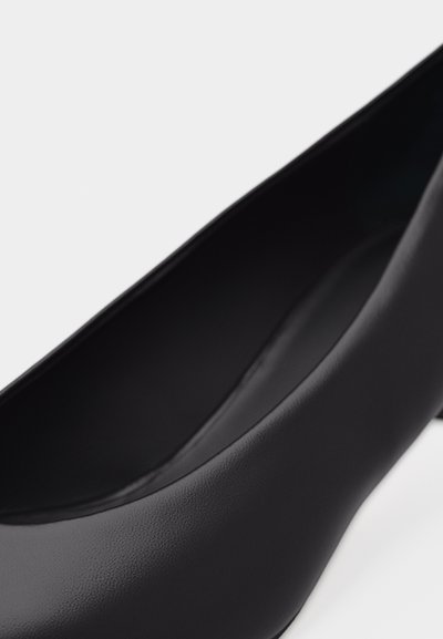 Κοντινή λήψη ενός μαύρου δερμάτινου παπουτσιού με ψηλό τακούνι, εστιάζοντας στην κυρτή εσωτε ρική πλευρά και την ομαλή υφή.