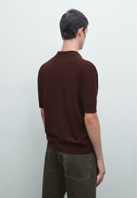 Burgundy kortärmad polo med klassisk krage och ribbade manschetter, ihop med mörkgröna jeans, sedd bakifrån.