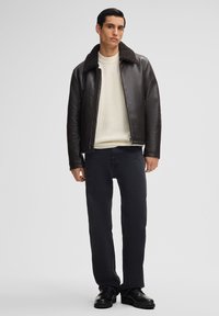 Bruine leren jas met een schapenvachtkraag, witte geribde trui, zwarte jeans en zwarte veterschoenen, staande tegen een effen achtergrond.