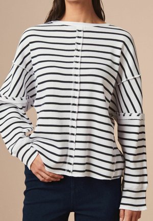 Nainen, jolla on valkoinen ribbineulottu pitkähihainen paita, jossa on mustat vaakaraitat ja keskellä pystysuora sauma, yhdistettynä tummiin housuihin.