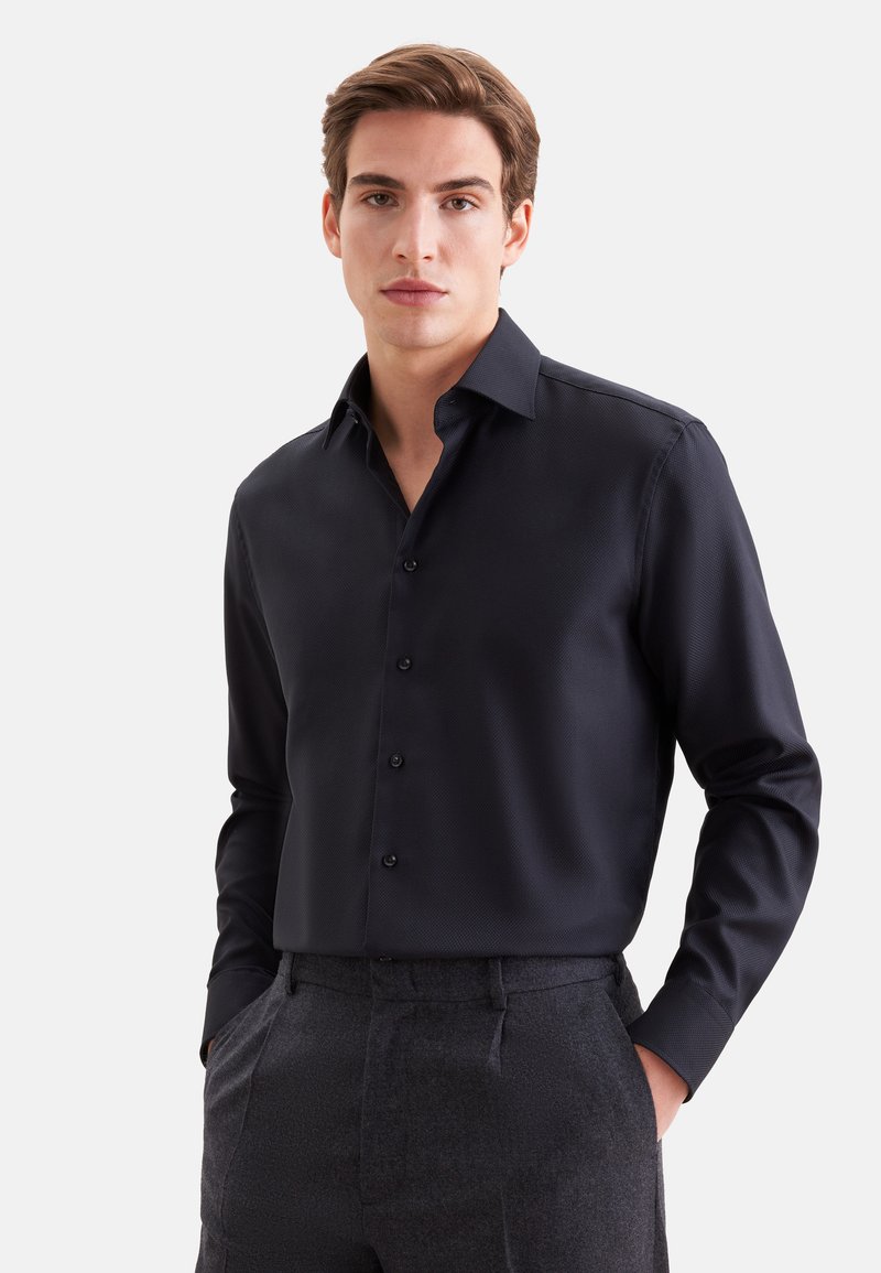Homme portant une chemise noire à manches longues boutonnée et un pantalon gris foncé, les mains dans les poches, sur un fond clair uni.
