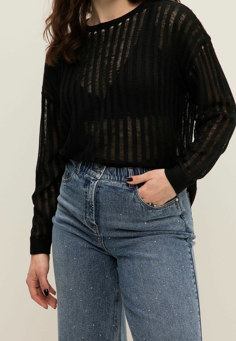 Maglione trasparente a righe nere con maniche lunghe, abbinato a jeans blu a vita alta con decorazioni in argento sparse.