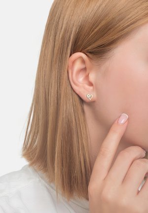 Goldene herzförmige Ohrstecker mit einem kleinen Edelstein, getragen an einem Ohr eines Models. Das Haar ist glatt und hellbraun, berührt die Schulter.