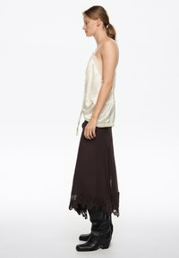 Kobieta w kremowej satynowej koszulce na ramiączkach, ciemnobrązowej spódnicy wykończonej koronką i czarnych, sięgających do kolan botkach, stojąca profilowo na białym tle.