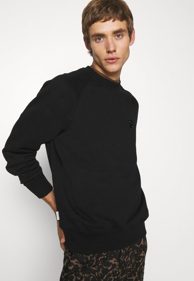 Sweatshirt noir avec des poignets et un col côtelés, comportant une petite patch logo noir sur la poitrine, porté sur un pantalon marron à motifs.