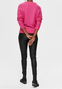 Model in een roze trui met opgerolde mouwen, met de rug naar ons gekeerd. Ze draagt zwarte leren broek en laarzen, met haar handen op de rug gekruist.