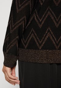 Pull en maille noire avec un motif zigzag métallisé doré, doté de poignets et d'un ourlet côtelés. La texture semble douce avec un effet scintillant.