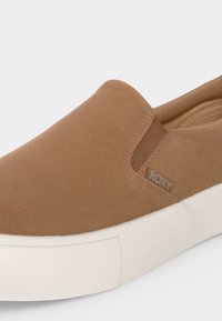 Chaussure slip-on beige avec semelle en caoutchouc blanche et empiècement latéral élastique, avec une petite étiquette en tissu avec le nom de la marque "Roxy."