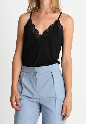 Nainen, jolla on päällään musta pitsireunainen toppi ja vaaleansiniset korkeavyötäröiset laskostetut housut, seisoo rennosti käsivarret alhaalla.