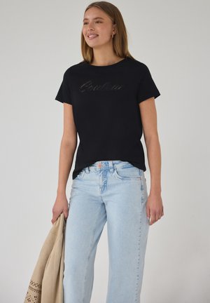 Jeune femme souriante, portant un t-shirt noir à manches courtes avec le texte « Couleur » et un jean bleu clair, tenant une veste beige dans la main gauche.
