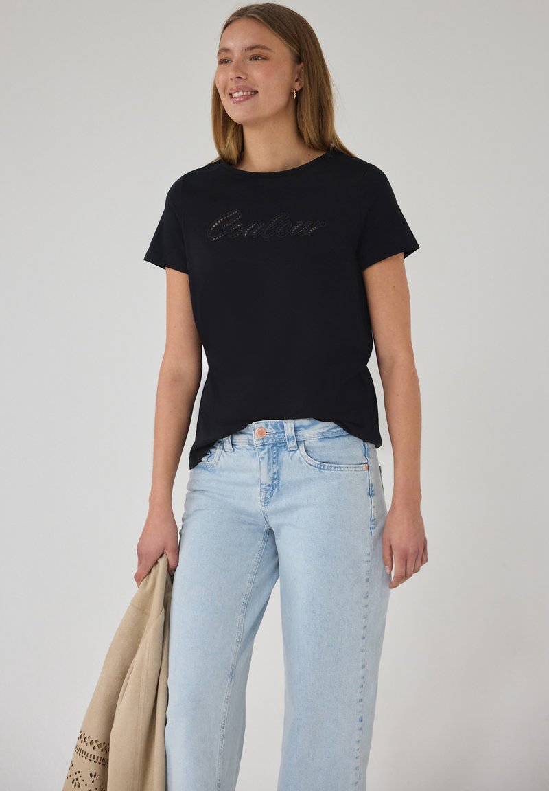 Jeune femme souriante, portant un t-shirt noir à manches courtes avec le texte « Couleur » et un jean bleu clair, tenant une veste beige dans la main gauche.