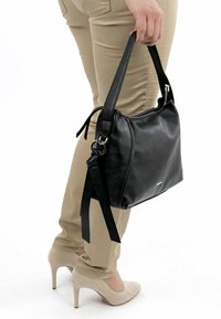 Musta nahkakäsilaukku, jossa on teksturoitu pinta, yksi olkahihna ja hopeinen vetoketjun yksityiskohta. Henkilöllä, joka käyttää vaaleita housuja ja korkokenkiä.