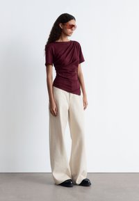 Weinrotes, gerafftes T-Shirt mit kurzen Ärmeln, kombiniert mit weiten beigen Hosen und schwarzen Schuhen. Das Model trägt orangefarbene Sonnenbrillen. Einfarbiger Hintergrund.