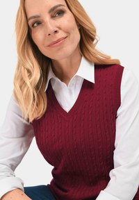 Bordeaux gebreid vest met een V-hals en een kabelpatroon, gelaagd over een witte blouse met kraag. Voorzien van ribgebreide randen en een mouwloze design.