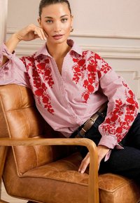 Blusa a righe rosse e bianche con ricami floreali, davanti con bottoni e maniche a sbuffo. Indossata con jeans scuri e una cintura marrone.