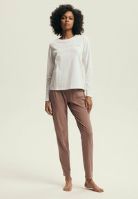 Biała bawełniana koszulka z długim rękawem z napisem "tylko dobre wibracje", połączona z miękkimi, jasnobrązowymi spodniami. Oba elementy są luźne i mają gładką teksturę.