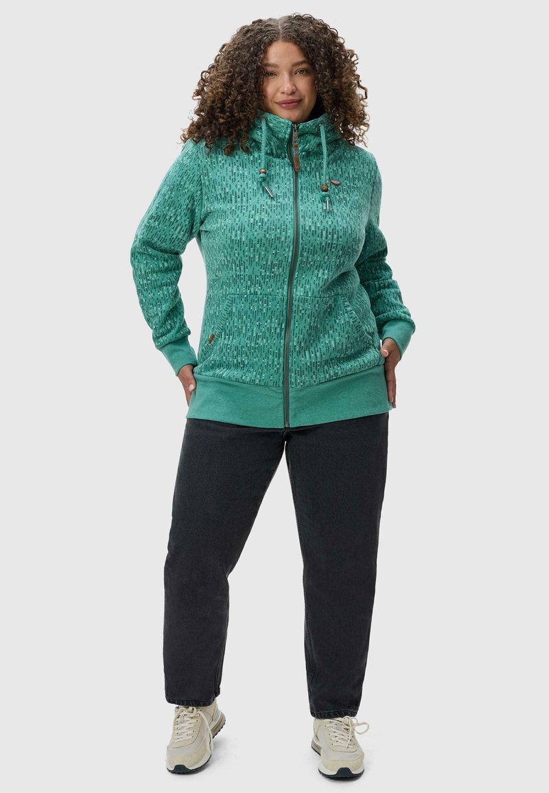 Hoodie zippé bleu sarcelle avec un motif texturé, des poignets élastiques et une capuche à cordon. Assorti avec un pantalon gris foncé et des baskets de couleur claire.
