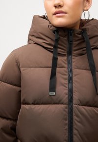 Pruun puffer-jope kõrge kraega, millel on eesne lukku, mustad tõmblukud ja logo silt rinnal. Sile tekstuur, tikitud disain.