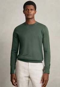 Dunkelgrüner Strickpullover mit rundem Halsauschnitt und langen Ärmeln. Der Pullover hat eine glatte Textur und sitzt auf Taillenhöhe, kombiniert mit hellen Hosen.