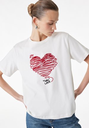 Kobieta z włosami związanymi w koczek, nosząca srebrne kolczyki koła i białą koszulkę z czerwonym bazgranym sercem oraz napisem „Endless Love”.