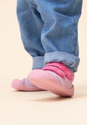 Light pink shoes with textured rubber sole and adjustable pink strap, paired with cuffed blue jeans. Background is neutral.