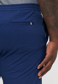 Mørkeblå bukser med elastisk talje, der har en baglomme med en flerfarvet stribet detalje. Stoffet ser glat og let ud.