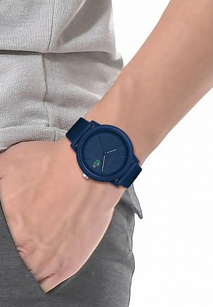 Reloj de pulsera azul marino con una esfera redonda, correa de goma texturizada y un pequeño logo de cocodrilo verde. Llevarlo en una mano en un entorno casual.