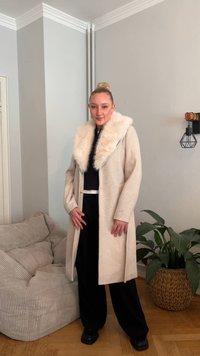 Casaco bege de lã com gola de pelo sintético, fecho de um botão e bolsos laterais. Combinado com calças pretas de perna larga e sapatos pretos robustos.