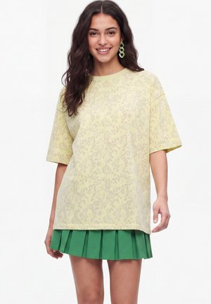 Ung kvinde smilende, iført en lys gul mønstret oversized T-shirt, grøn plisseret nederdel og grønne geometriske øreringe, stående mod hvid baggrund.