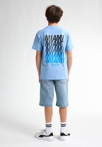 T-shirt bleu à manches courtes avec le texte "MIAMI" empilé en noir et bleu, associé à un short en denim clair et des baskets blanches.