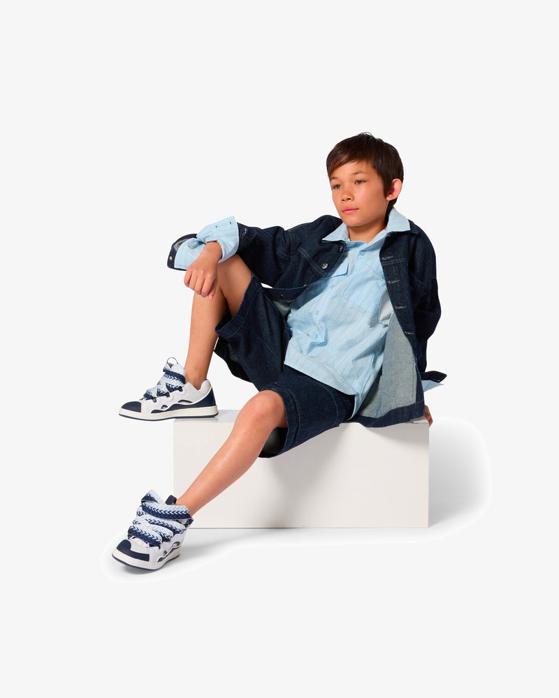 Veste en denim, chemise rayée bleu clair, short en denim, baskets blanches avec des accents bleus. Modèle assis sur une boîte blanche, pose décontractée.