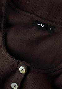 Een bruin geribbeld stoffen topje met een ronde halslijn, voorzien van twee parelachtige knopen en een label met de tekst "LMTD 14 S." Een gestructureerd ontwerp detail is zichtbaar.