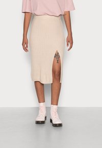 Kráčející midi sukně v béžové barvě s žebrovanou strukturou a bočním rozparkem; kombinována se světle růžovými šněrovacími botami. Jednoduchý a přiléhavý design.