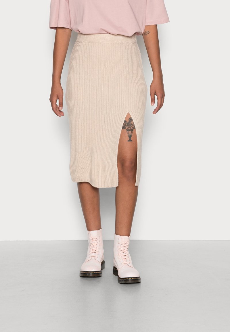 Kráčející midi sukně v béžové barvě s žebrovanou strukturou a bočním rozparkem; kombinována se světle růžovými šněrovacími botami. Jednoduchý a přiléhavý design.