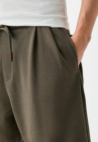 Tmavě zelené vroubkované kalhoty se bočními kapsami a pasem se stahovací šňůrkou. Mají strukturovanou tkaninu a dřevěný detail na stahovací šňůrce.