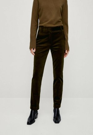 Persoon die donker olijfgroene velours rechte broek draagt, een bruine top met lange mouwen met verlengde manchetten en zwarte enkellaarsjes, staand op een witte vloer.