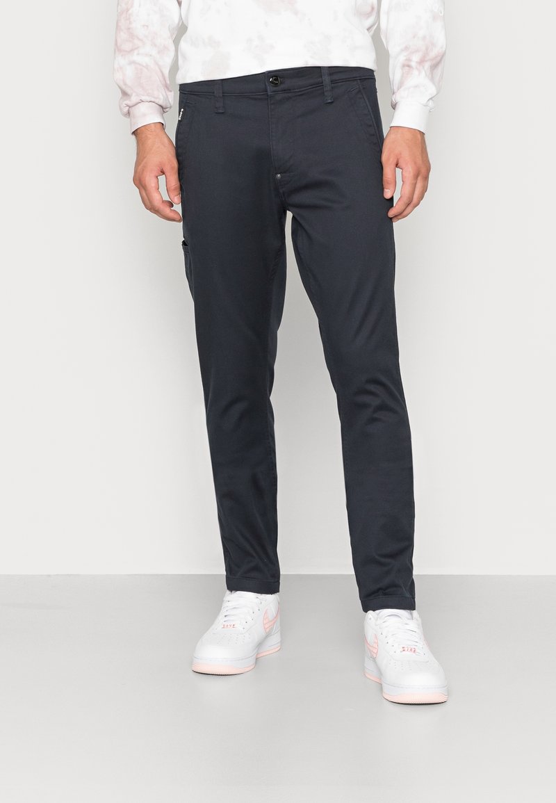 Chinos bleu foncé à coupe slim, avec un devant plat, des poches latérales et un détail zippé sur la gauche. Ils sont assortis à des baskets blanches.