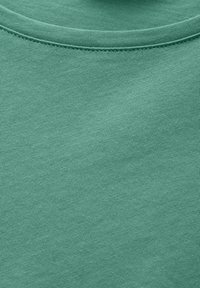 Vihreä puuvillapaita, jonka kangas on sileä, esittelee ribbausta kaula-aukossa ja tasaisesti ommeltuja saumoja, ja sen väri on tasainen.