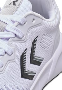 Witte sportschoen met zwart dubbel chevron-logo, witte veters en een gestructureerd stoffen bovenwerk in close-up.