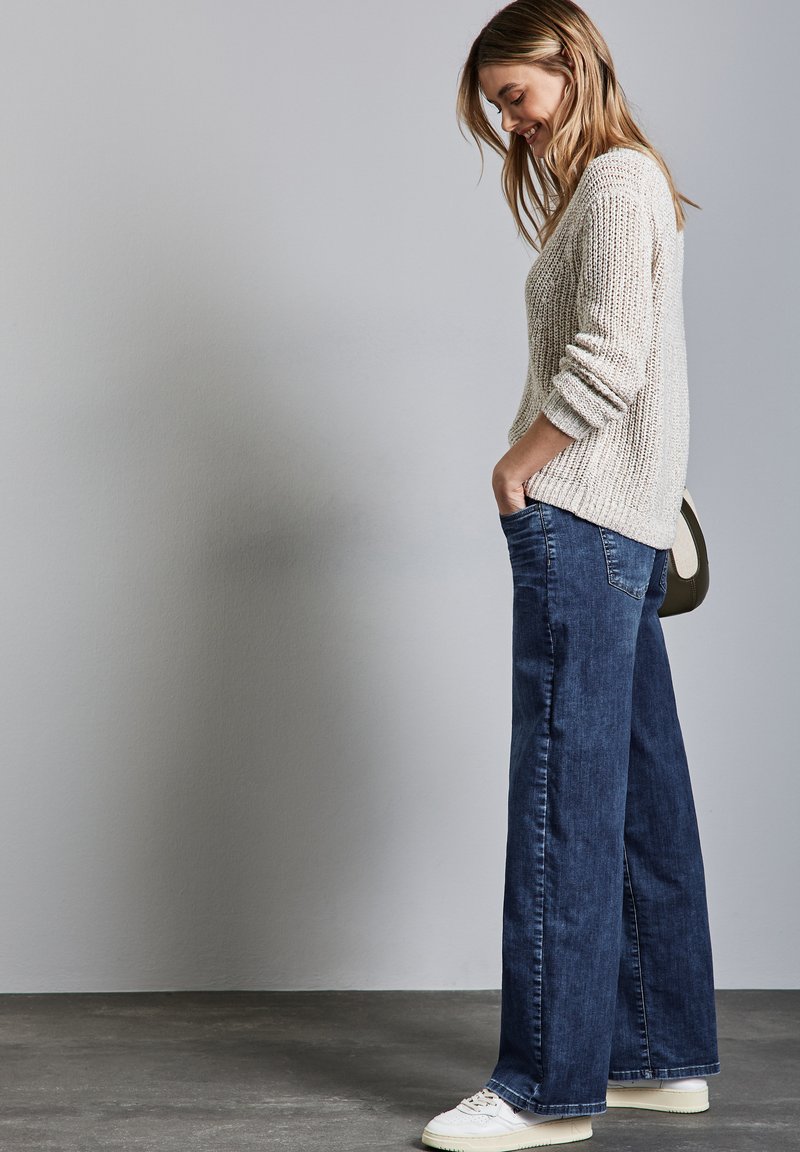 Suéter de punto texturizado en gris claro combinado con jeans de mezclilla oscuros de tiro alto y pierna ancha, junto con zapatillas blancas. Conjunto sencillo y casual.