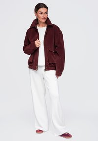 Femme portant une veste rouge foncé sur une chemise blanche avec un pantalon blanc large et des baskets rouge foncé, debout devant un fond clair.