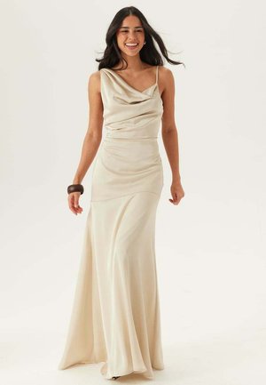 Kvinde smilende og gående fremad iført en lang, cremefarvet satin kjole med en tynd strop og draperet udskæring, med et mørkt armbånd på venstre håndled.