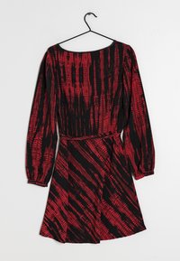 Röd och svart långärmad klänning med en strukturerad mönster, åtsittande midja och utsvängd kjol. Hänger på en svart krok mot en vit bakgrund.