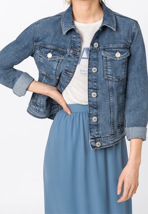 Femme portant une veste en jean bleue à manches retroussées sur un t-shirt blanc à motif graphique et une jupe bleue taille haute, main sur la hanche.