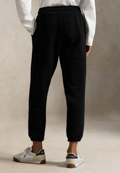 Melnas sporta bikses ar elastīgu jostasvietu, gali sašaurināti pie potītēm. Valkātas ar baltām sporta kurpēm, kurās ir tumši zils akcents un logo.